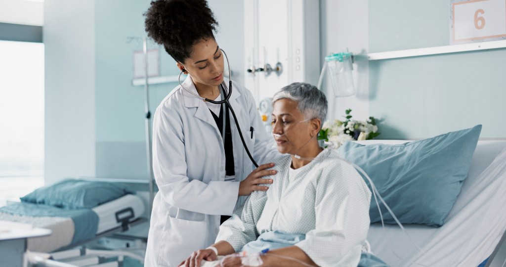 A female doctor examines an elderly female patient with a stethoscope in a hospital room.