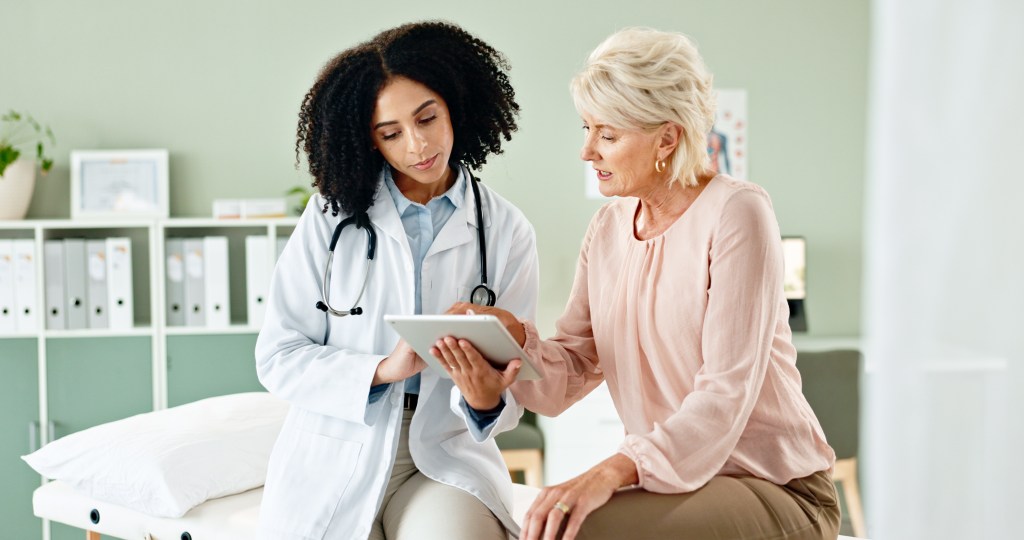 A doctor and an elderly female patient look at a tablet together in a clinic.