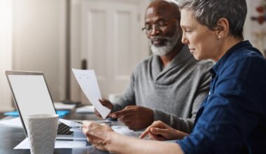 A senior Black man wearing glasses and a gray sweater holds a document while an older white woman in a blue shirt points to papers on a desk next to a laptop and a textured white coffee mug. They are both focused on the documents, appearing to discuss finances in a well-lit home setting.