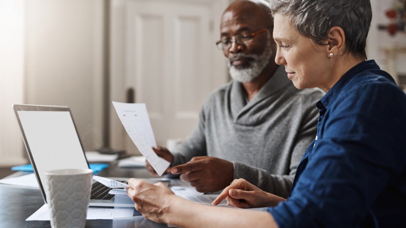 A senior Black man wearing glasses and a gray sweater holds a document while an older white woman in a blue shirt points to papers on a desk next to a laptop and a textured white coffee mug. They are both focused on the documents, appearing to discuss finances in a well-lit home setting.