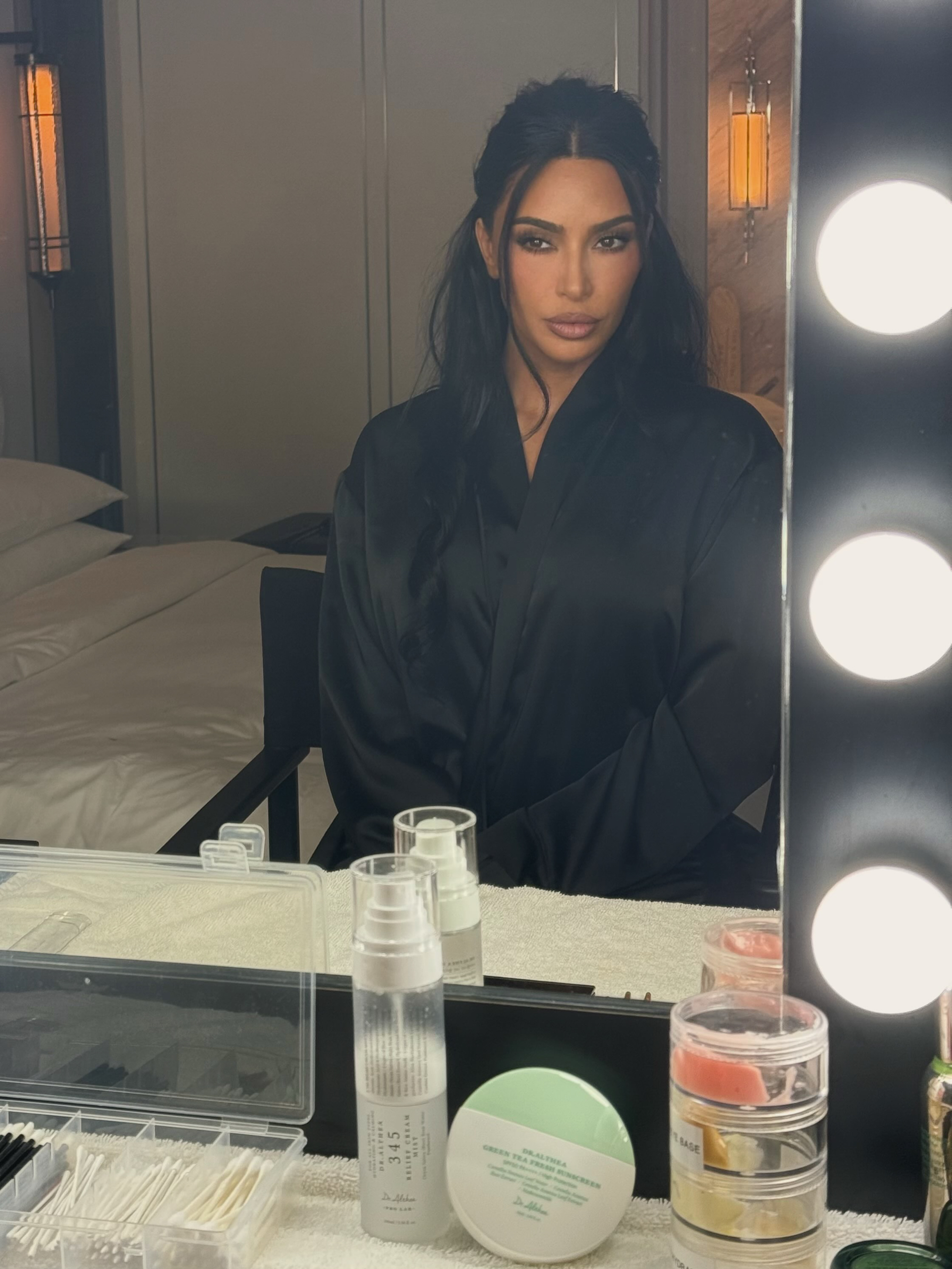 Kim Kardashian wearing a black robe while looking into a lighted vanity mirror.