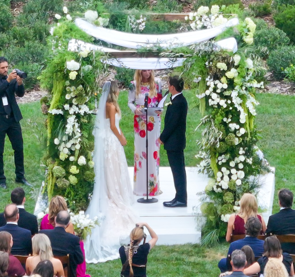 Matthew Lowe and Emily Markus exchange vows under a chuppah decorated with white flowers and greenery in Santa Barbara, California, with an officiant and seated guests.
