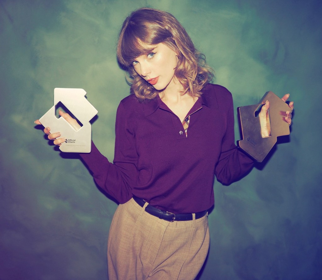 Taylor Swift holds two arrow-shaped Official Charts awards in front of a green backdrop.