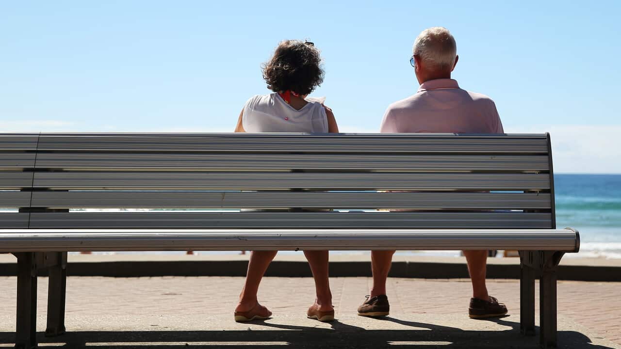 An older couple sit on a bench facing the ocean as seen from behind.