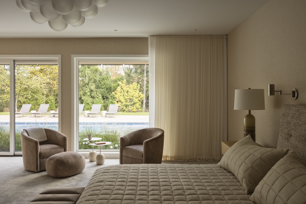 A bedroom with a quilted bed, two armchairs, an ottoman, and large windows overlooking a swimming pool.