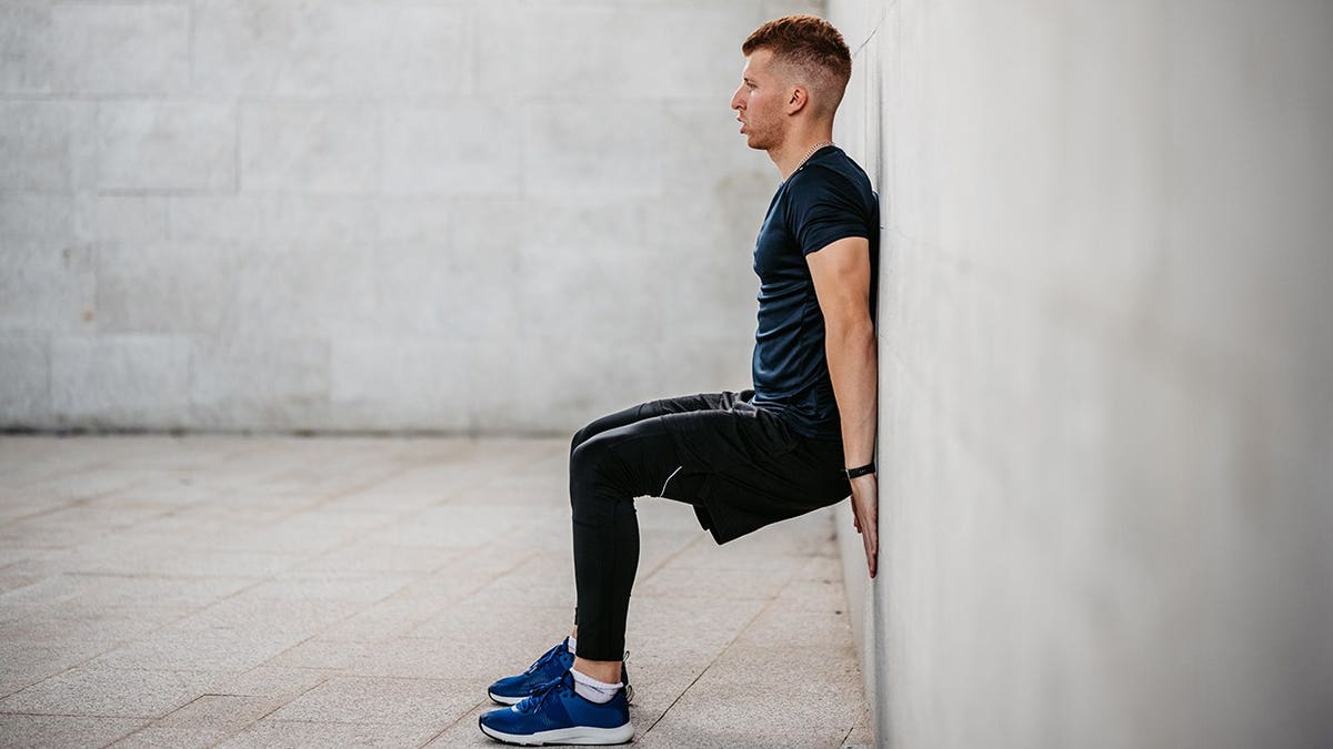 Young man performing a wall sit exercise outdoors against a wall.