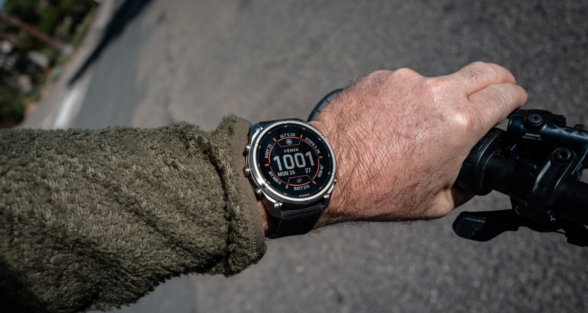 A cyclist's wrist in an olive fleece sleeve wearing a Garmin fenix watch with a silver bezel, its face showing 10:01, while the hand grips a bike handlebar on an asphalt road.