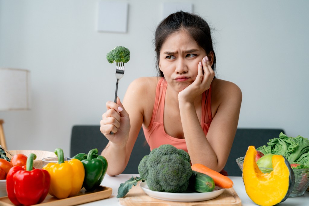 A young Asian woman makes a disgusted face while holding a fork with broccoli and looking at vegetables on a table.