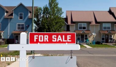 A red sign reading "For Sale" hangs on a residential street, in front of two single family homes.