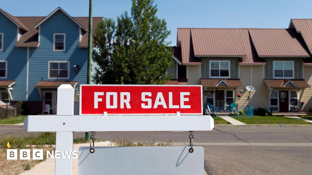 A red sign reading "For Sale" hangs on a residential street, in front of two single family homes.