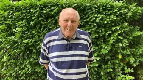 A man wearing a t-shirt with stripes, stands outside in front of a large green bush. His hands are tucked behind him.