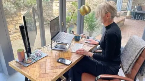 A woman sitting at a desk in a home office. She is wearing a black turtleneck and blonde hair.
On the desk are several pairs of glasses, a monitor, a calculator, a laptop on a stand, a phone and a mug.