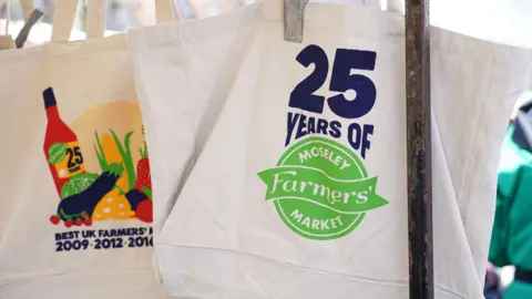 Moseley Farmers' Market A white reusable shopping bag with "25 years of Moseley Farmers' Market" written on the side of it in navy blue above a green logo.
