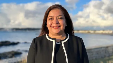 BBC Munazza Malik, with dark brown hair, wearing a black blazer and top with a white collar piping around the sides. She is standing in front of a beach.