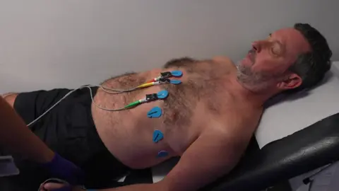 Chris is lying on a hospital bed, with six blue medical pads strapped to his chest, three have green, yellow and red wires coming out of them, connected to something out of shot.