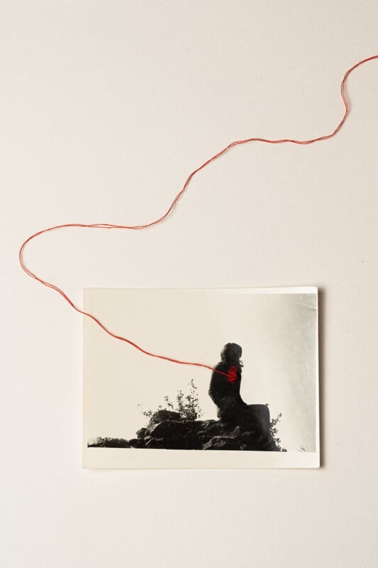 A black-and-white photo of a person sitting on a rock is placed on a white surface. A red string is attached to the photo, appearing to connect to the person's chest and extending outward across the background.