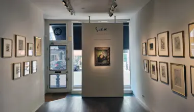 A spacious art gallery with framed artworks displayed on white walls. A central partition features a highlighted piece, with large windows letting in natural light.