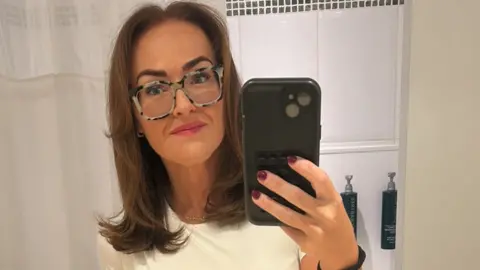 Emilly Murray @itsanewmeemj A woman with straight brown hair, a white t-shirt, patterned glasses and pink lipstick takes a selfie in the mirror with a phone