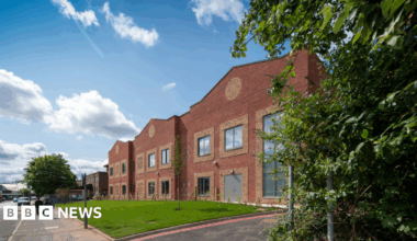 A long two-storey red brick hospital building pictured from the street. There is a grass area alongside, with people walking past.