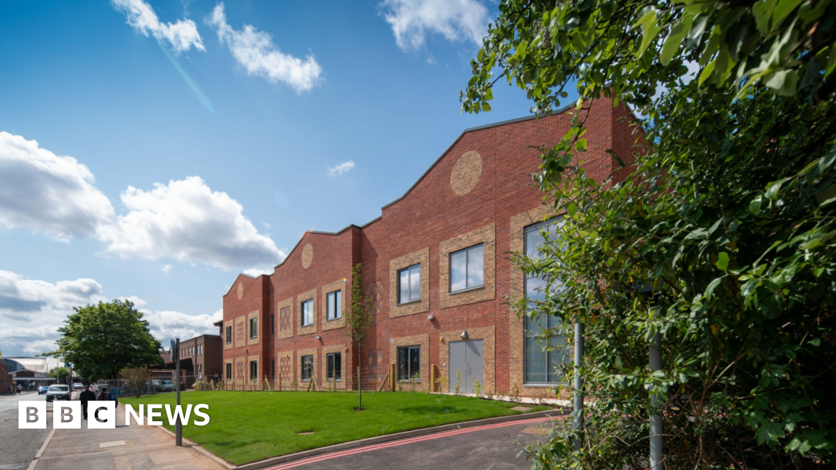 A long two-storey red brick hospital building pictured from the street. There is a grass area alongside, with people walking past.