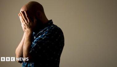 A man wearing a blue-checked shirt with his face buried in his hands