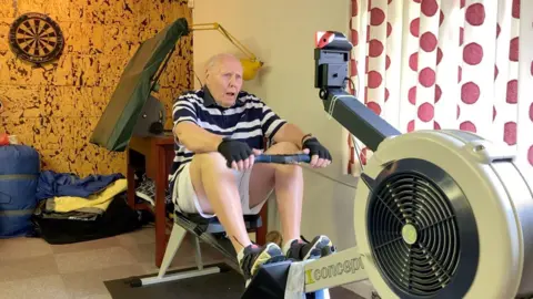 BBC A man wearing a t-shirt with stripes and shorts on, sits on a rowing machine in a home gym. 