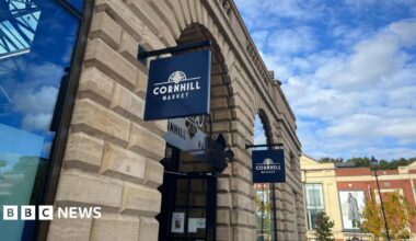 An exterior view of Cornhill Market showing a modern building with navy signs hanging above the arched entrance reading 'Cornhill Market'. The sky is blue with light clouds.