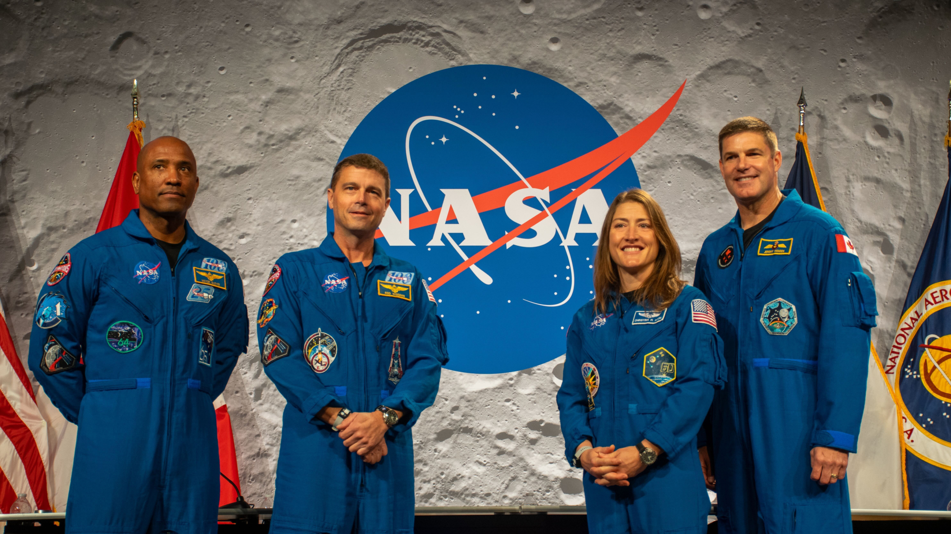 Four people wearing blue jumpsuits stand next to each other in front of a desk with a NASA logo behind them