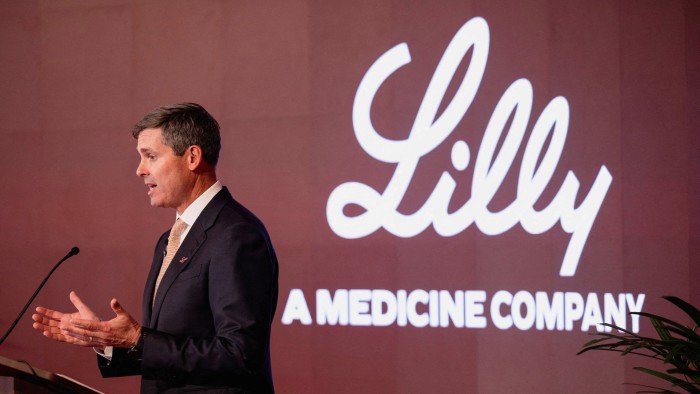Dave Ricks speaks at a podium in front of a screen displaying the Lilly logo and slogan
