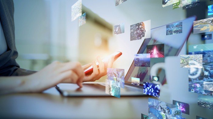 A person uses a smartphone and laptop surrounded by floating digital images,