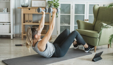 Woman performs sit-ups with dumbbells at home, with her feet hooked under a green armchair