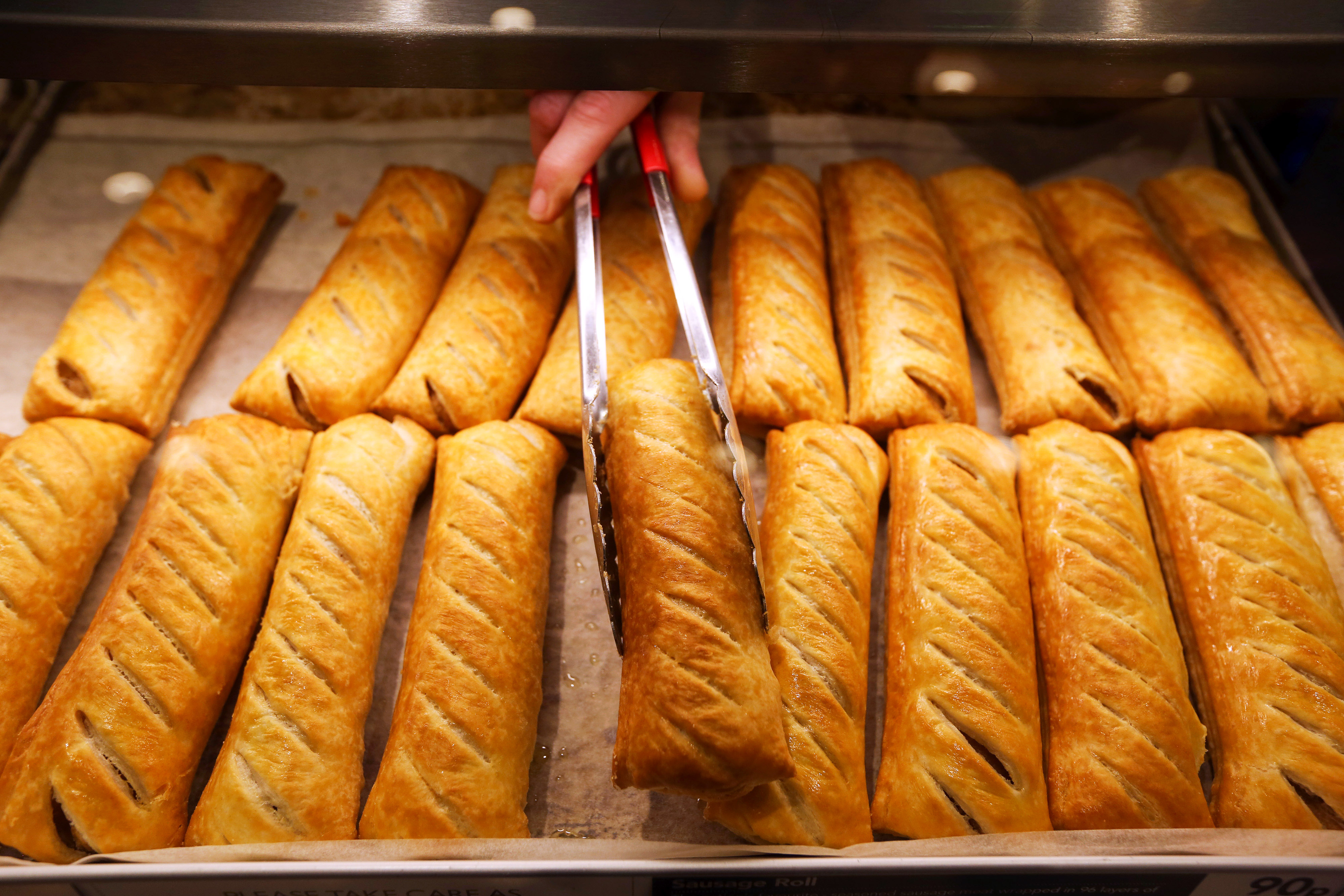 A hand using tongs to select a sausage roll from a display of many at a bakery.