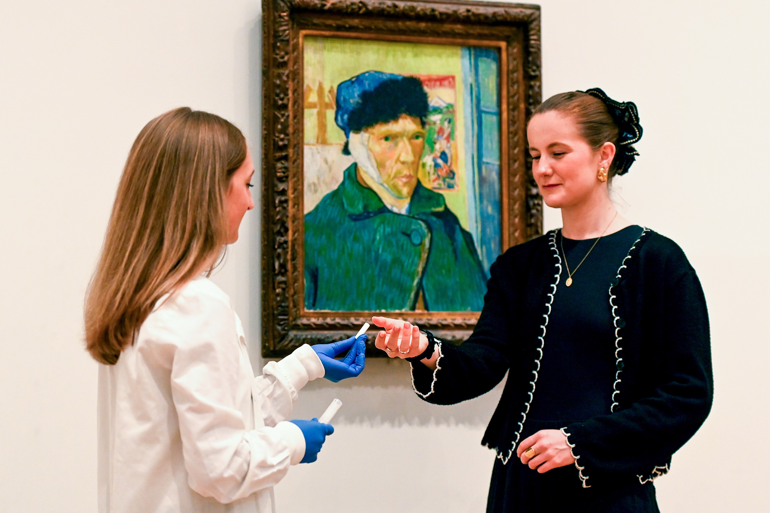 A researcher hands a participant a sample in front of Van Gogh's "Self-Portrait with Bandaged Ear" at a gallery.