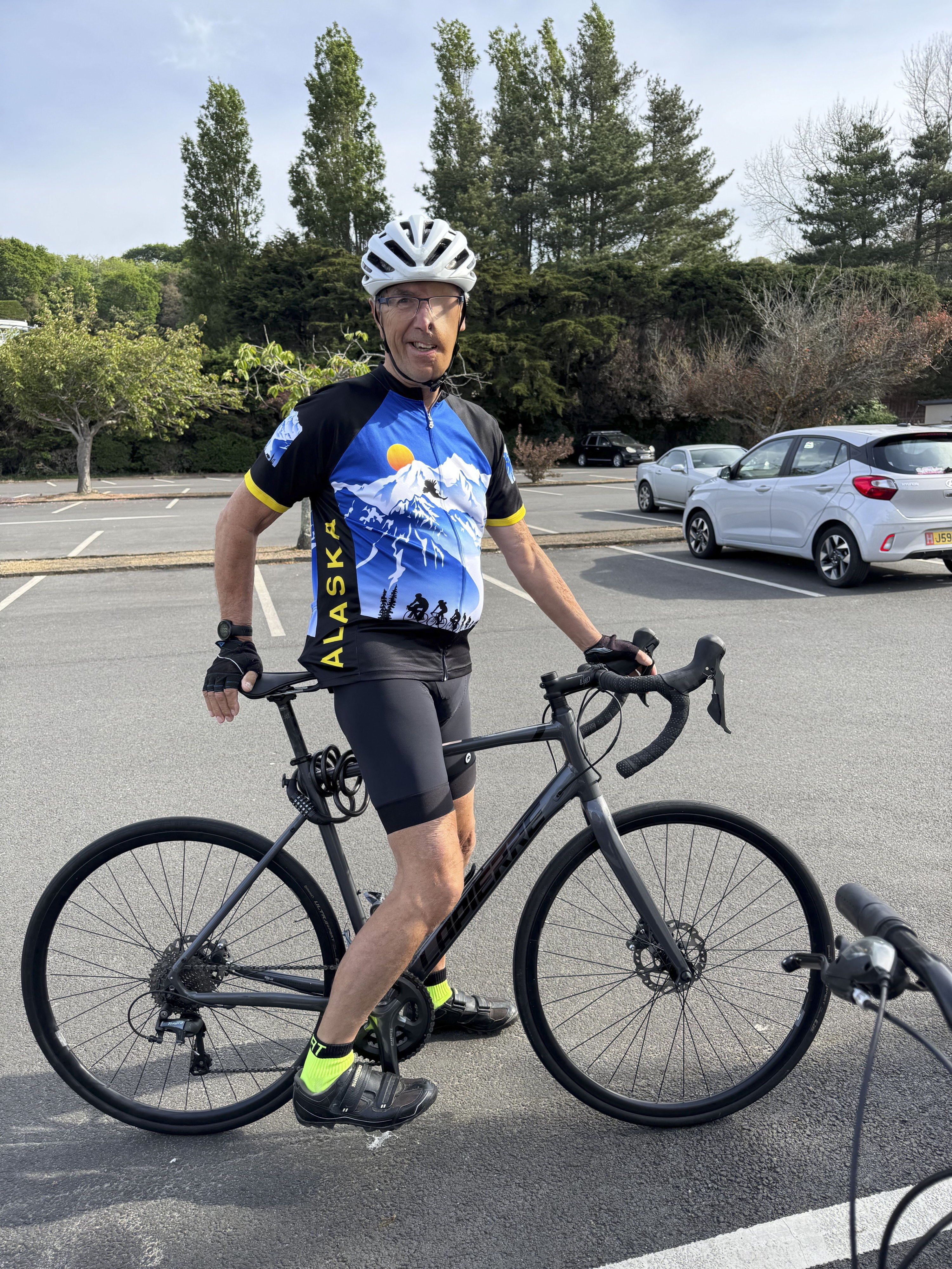 Dave Richards, 75, wearing a blue and black cycling jersey with "ALASKA" written on the side, standing next to his bike in a parking lot.