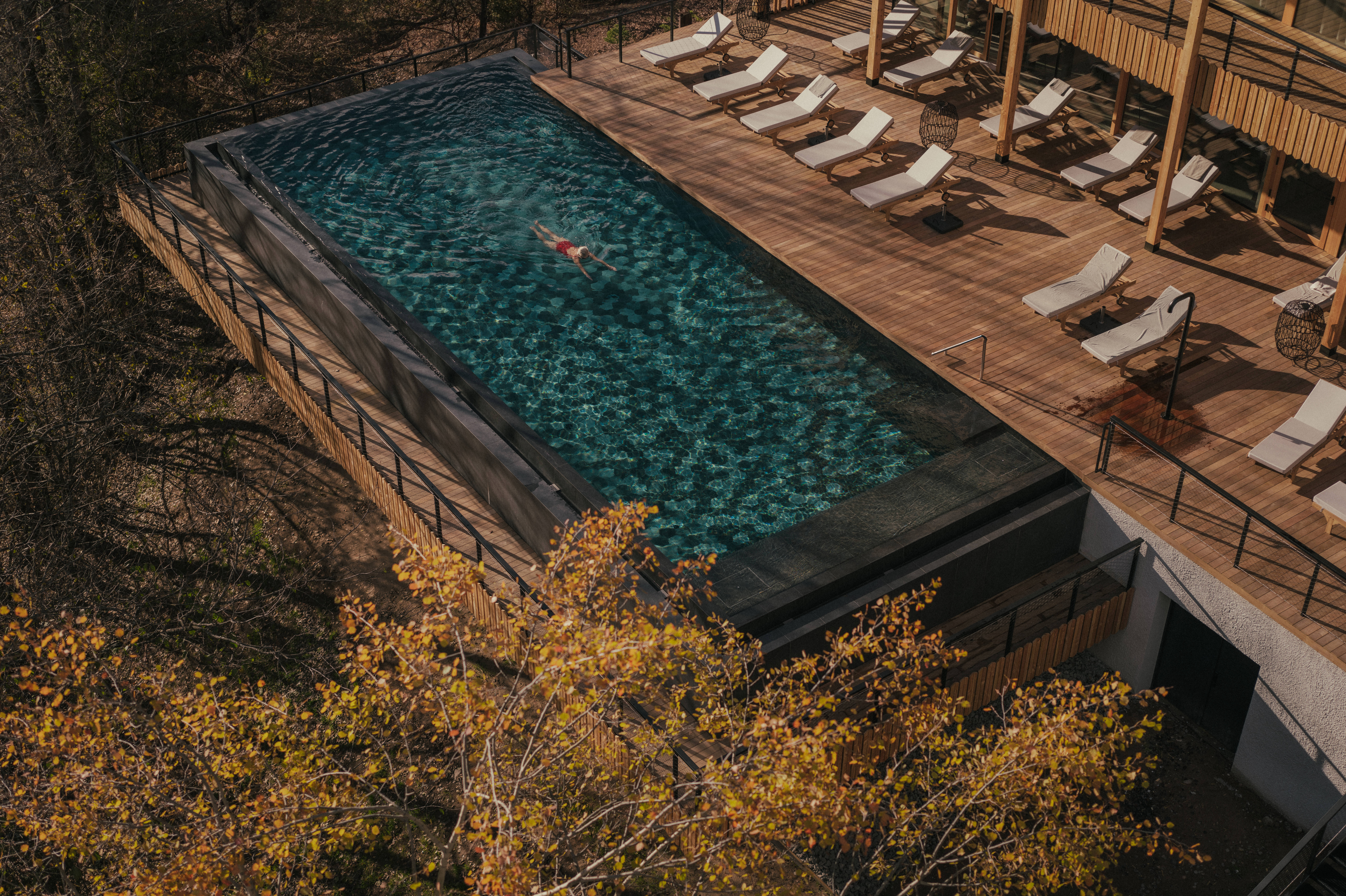An aerial view of a woman swimming in a pool next to a wooden deck with lounge chairs.