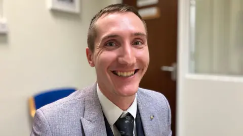 A man with short brown hair, in a grey suit with a black tie and waistcoat.