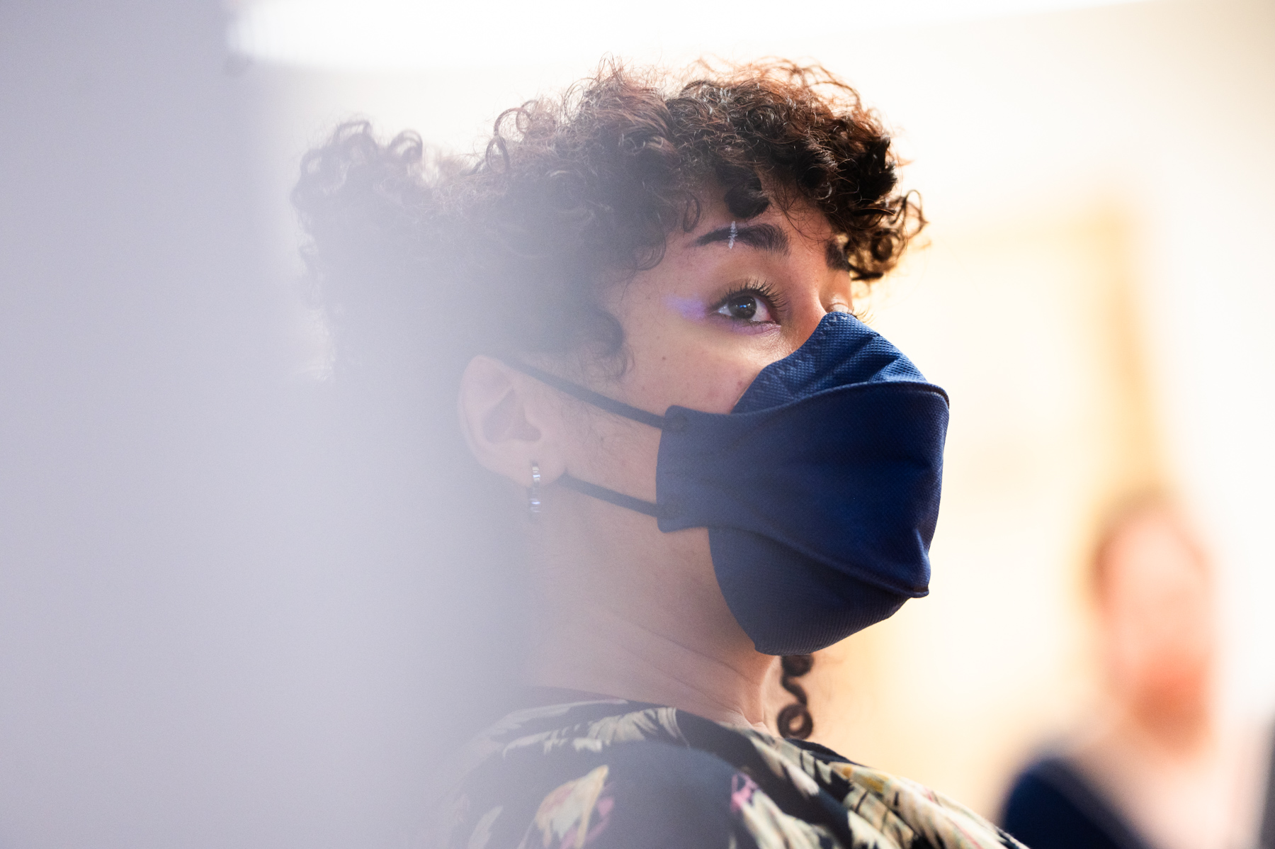 A person with curly hair and floral clothing wears a dark blue face mask, looking slightly upward. The background is softly blurred.
