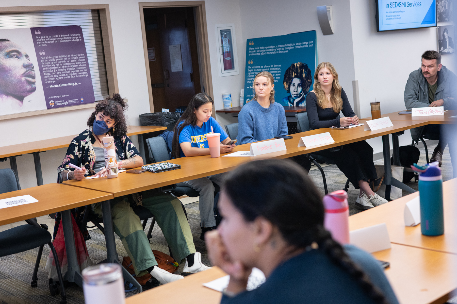A group of people sit at tables in a meeting room, listening attentively. Nameplates, notebooks, and water bottles are on the tables. A poster and a TV screen are visible in the background.