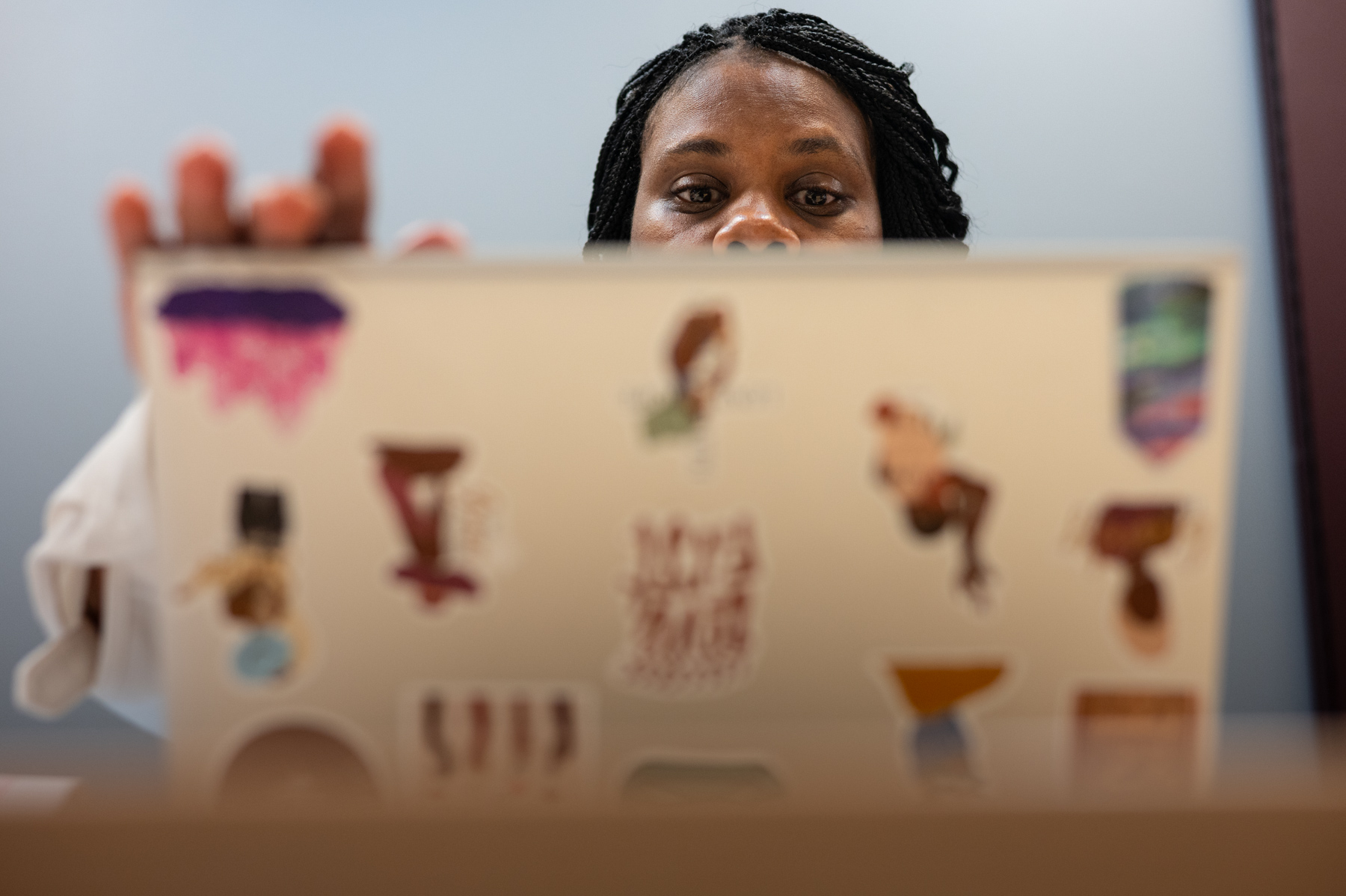 A person with braided hair works on a laptop decorated with various colorful stickers, viewed from the front of the laptop.