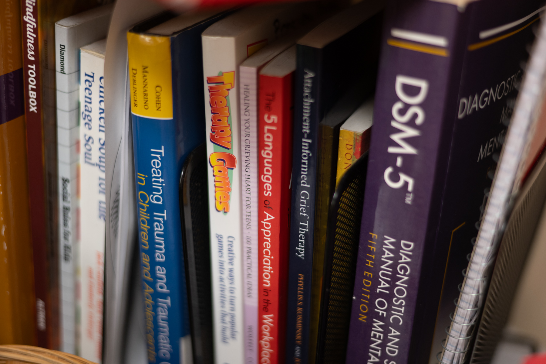 A close-up of several therapy and mental health books on a bookshelf, including titles like DSM-5, Therapy Games, and Treating Trauma and Traumatic Grief in Children.