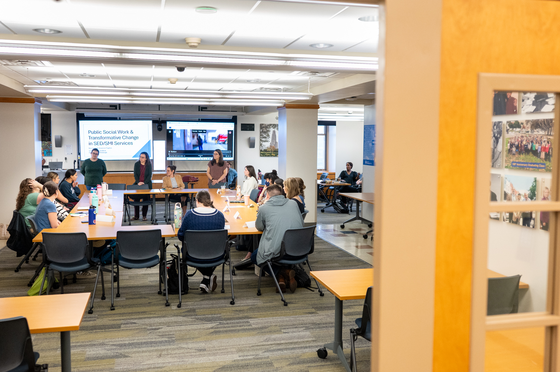 A classroom with people seated around tables, four presenters standing at the front, and a screen displaying "Public Social Work & Transformative Change in SED/SMI Services.