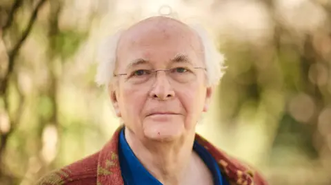 Sam Allard Philip Pullman with glasses in a brown & beige cardigan and blue shirt against trees and leaves background
