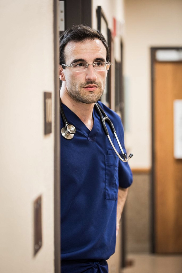Matt Heinz stands between a doorway and a hallway at a doctor's office, wearing dark blue scrubs and a stethoscope