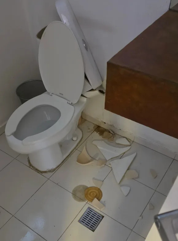 Toilet with lid up, surrounded by broken ceramic pieces on tiled floor; signs of a recent accident