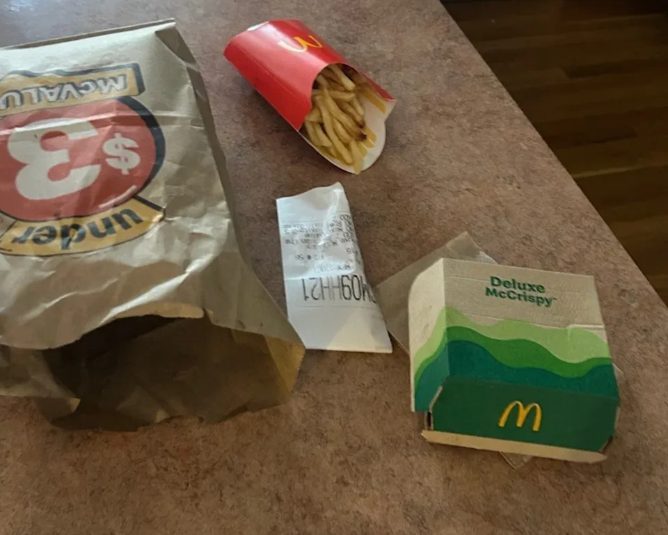 Fast food on a table, including a paper bag, fries, a receipt, and a Deluxe McCrispy box from McDonald's