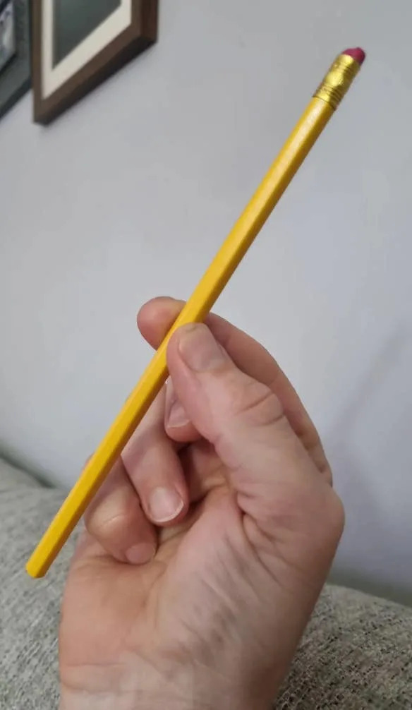 Hand holding a yellow pencil with an eraser, angled slightly upward. Frame pictures are blurred in the background