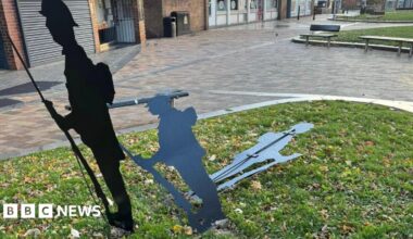 Three black metal silhouette sculptures depicting soldiers. One figure holding a rifle with a bayonet is standing upright while the second figure is bent lower and the third one near the ground. They are placed on a small grassy area with scattered autumn leaves, surrounded by a paved pedestrian space.