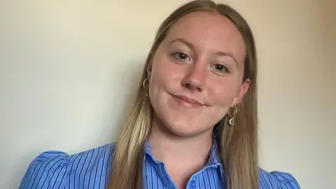 Ashleigh Sadler A woman with long blonde hair is wearing a striped blue blouse and smiling. There is a white wall behind her.
