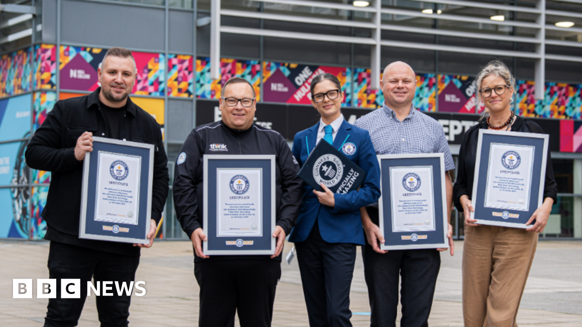Three men and two women stand side by side in front of a large, modern building with colourful designs on the exterior. Each person is holding a framed certificate from Guinness World Records, which features a blue and white design with the official logo at the top. They are facing the camera. The award reads: "The most users in a mental health lesson was achieved by Think Mental Health, Think Cloud, Hull College, Smith & Nephew (all UK) in Hull, UK, on 10 October 2025."