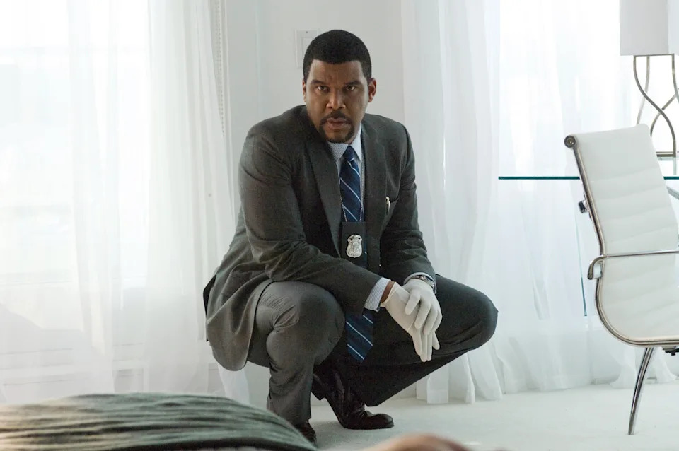 Tyler Perry in a suit, crouching, wearing white gloves, appears focused in a modern, bright room